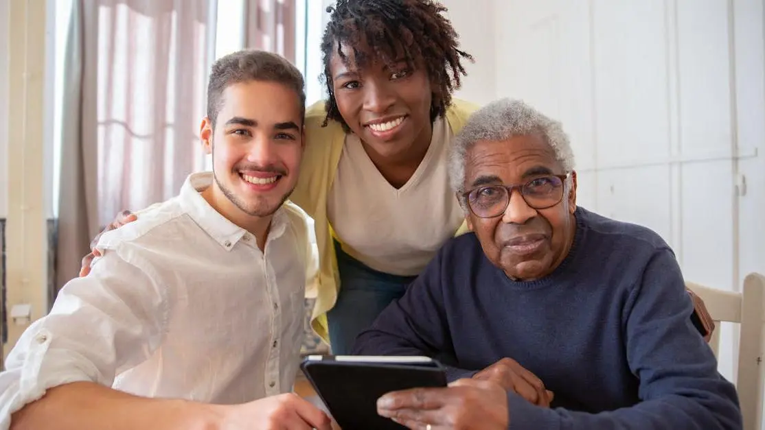 Volunteers at senior home helping elderly man use a tablet, smiling in a cozy living room